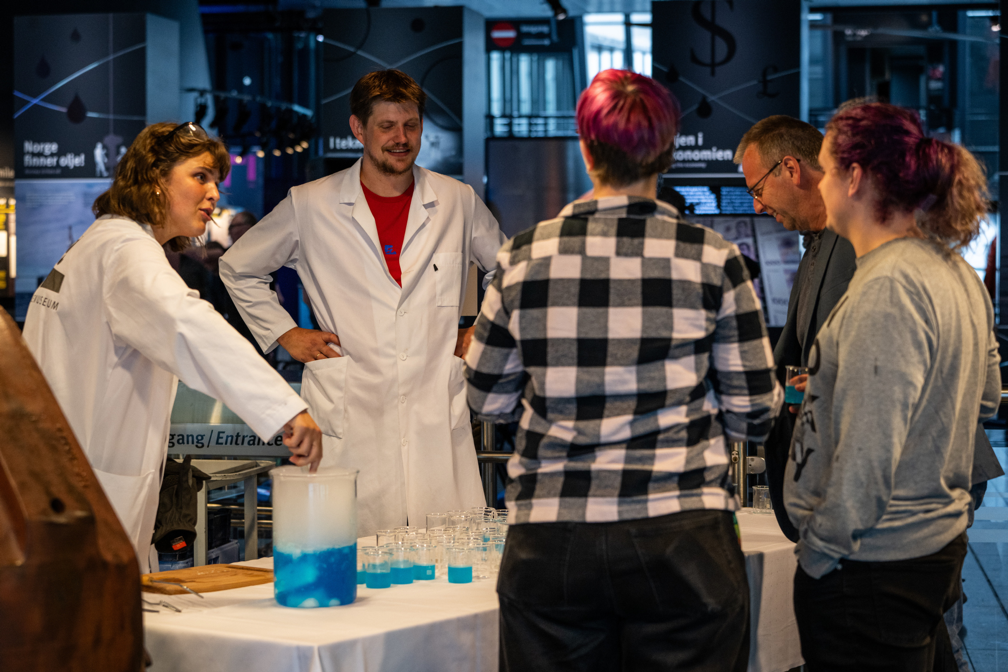 To personer i hvite labbfrakker serverer blå drikke i reagenskopper til publikummere