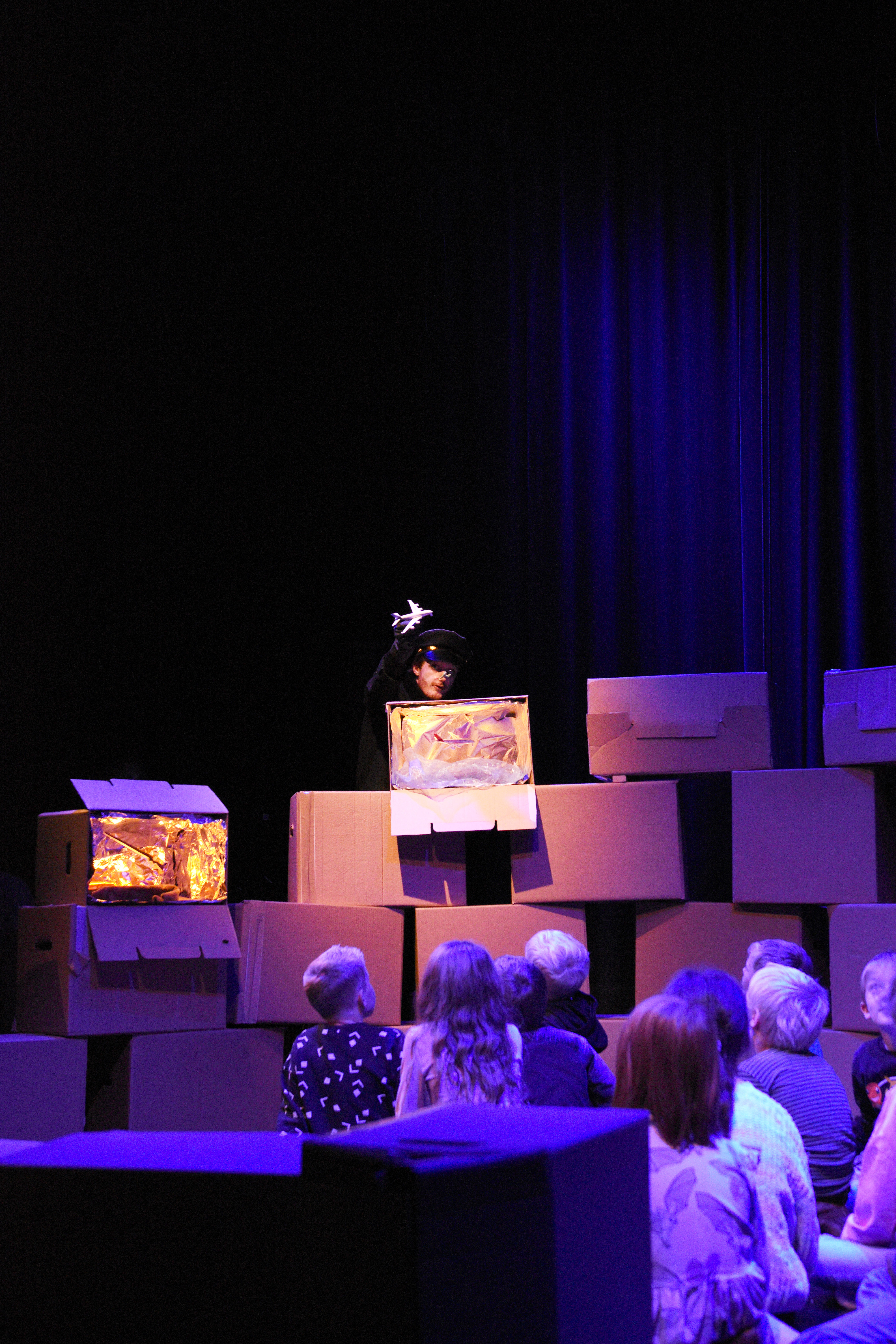 En skuespiller titter fram fra noen pappesker på en scene. Foran i bildet sitter en gruppe barn.