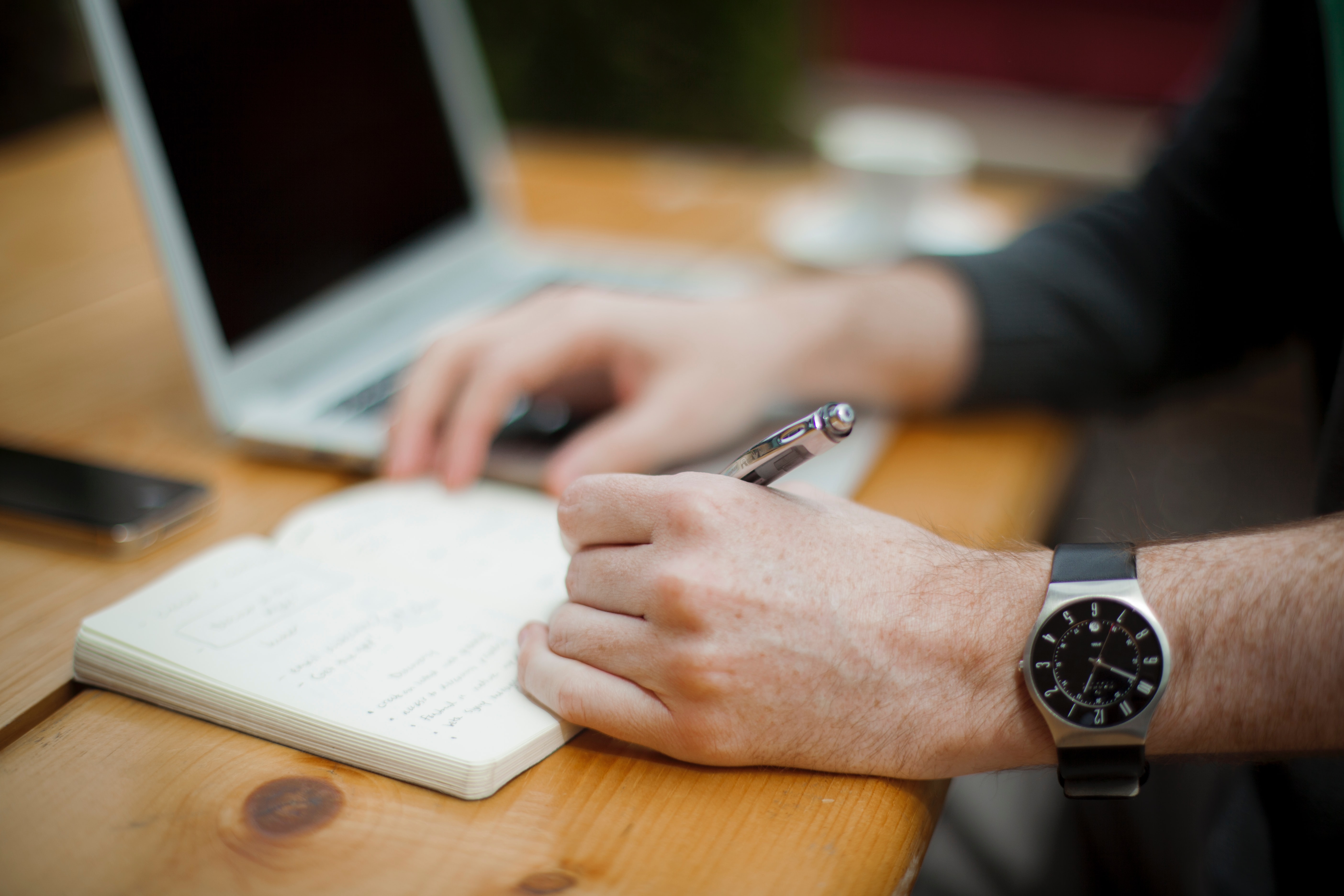 A man with a black watch is writing something i a notebook with his laptop beside him by Alejandro escamilla