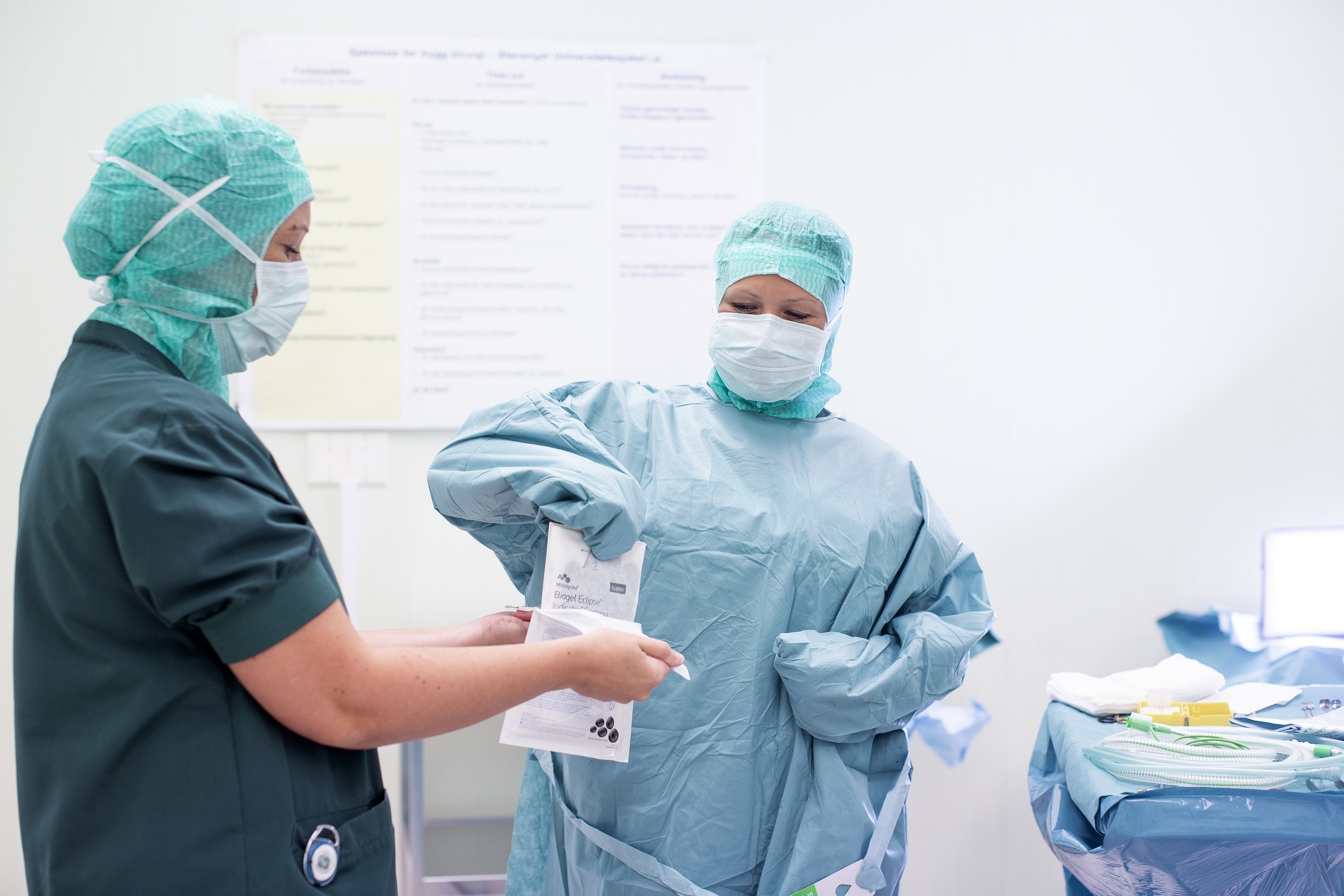 To kvinnelige operasjonssykepleiere i uniform på operasjonsstue. En av dem er sterilkledd og får assistanse til å ta på sterile hansker.