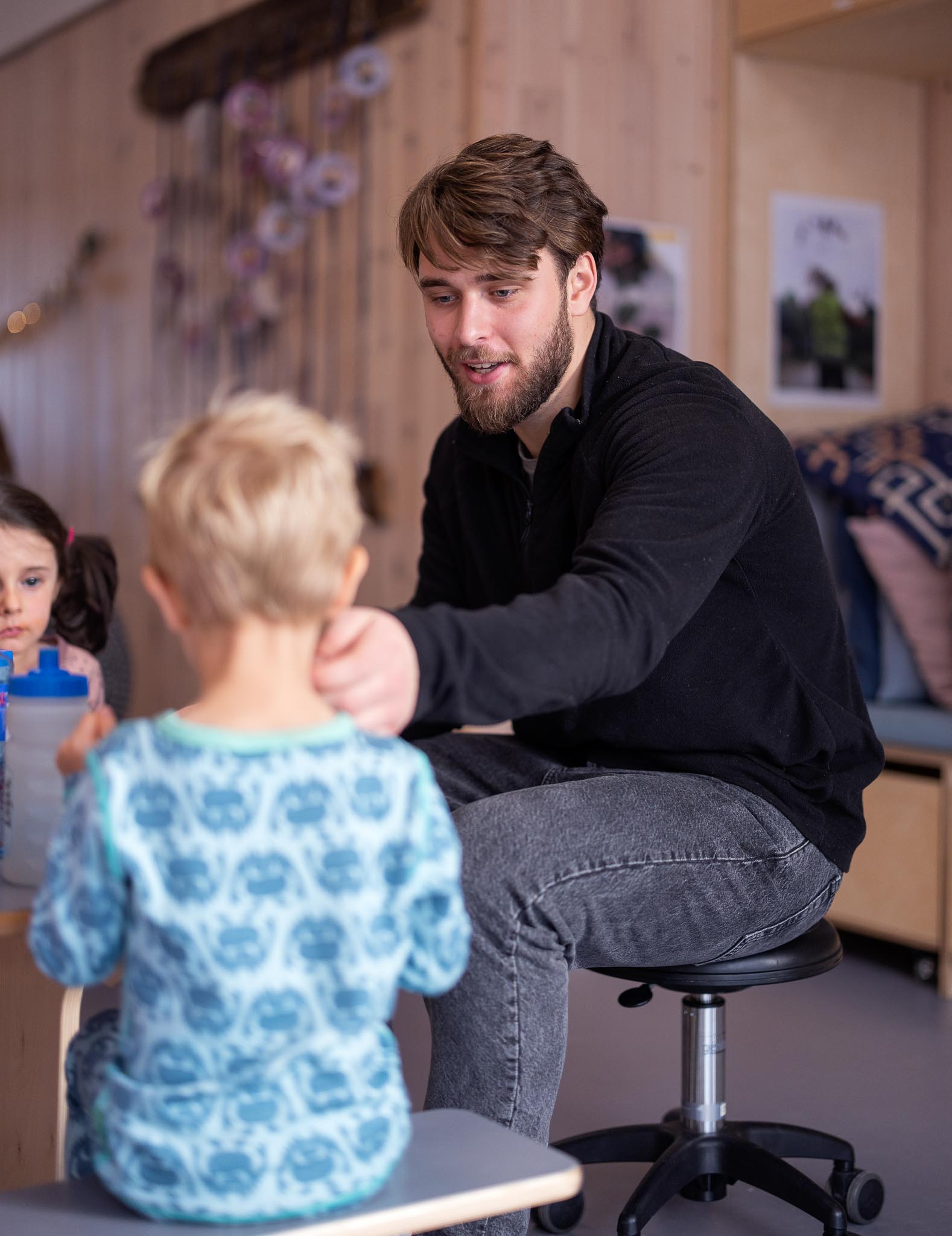 Ung mannlig barnehagelærer har på seg svart genser og jeans, sitter på en stol og hjelper et barn som sitter med ryggen til. 