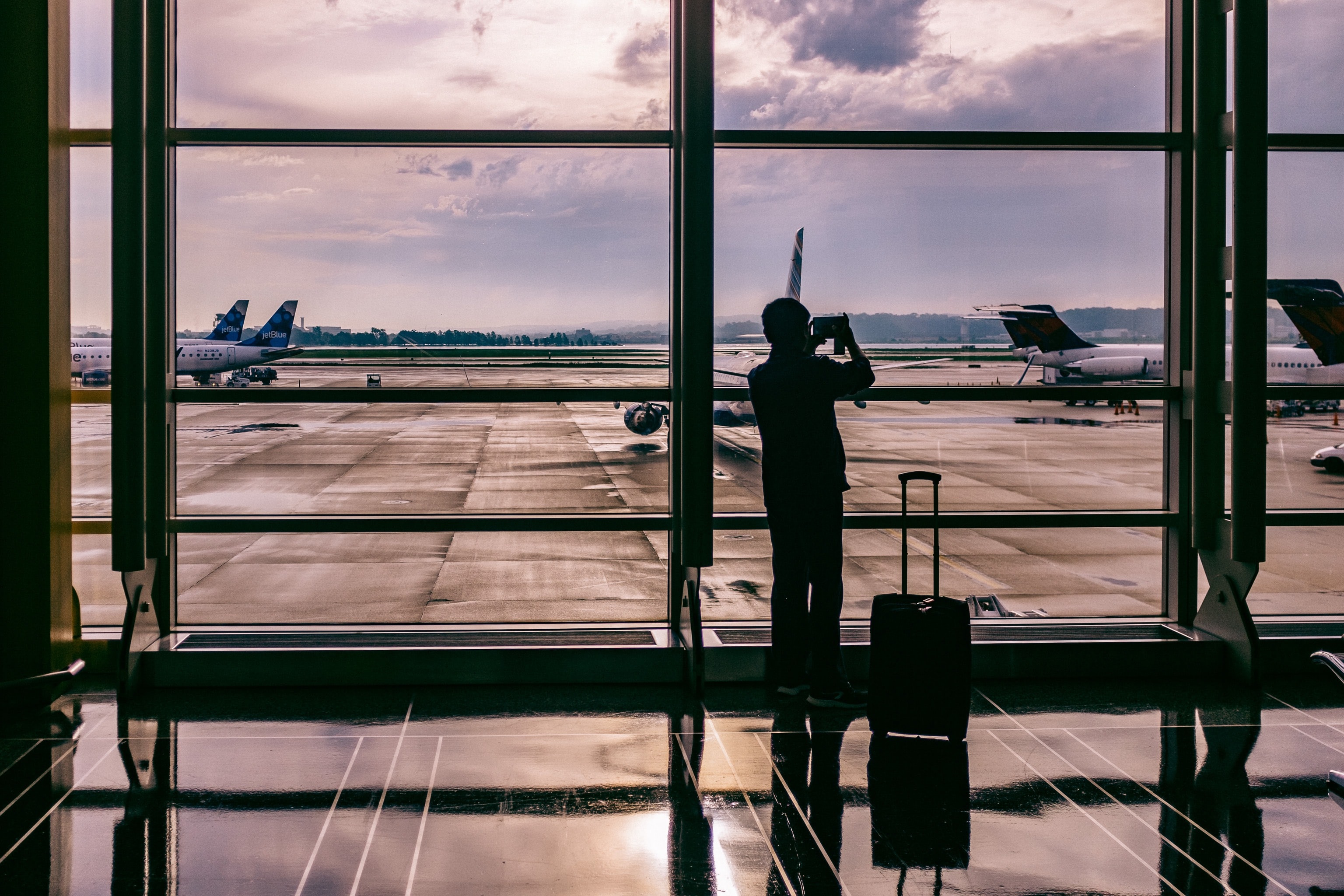 a silhouette of a man using his phone at an airport. the man is standing beside his suitcase. 