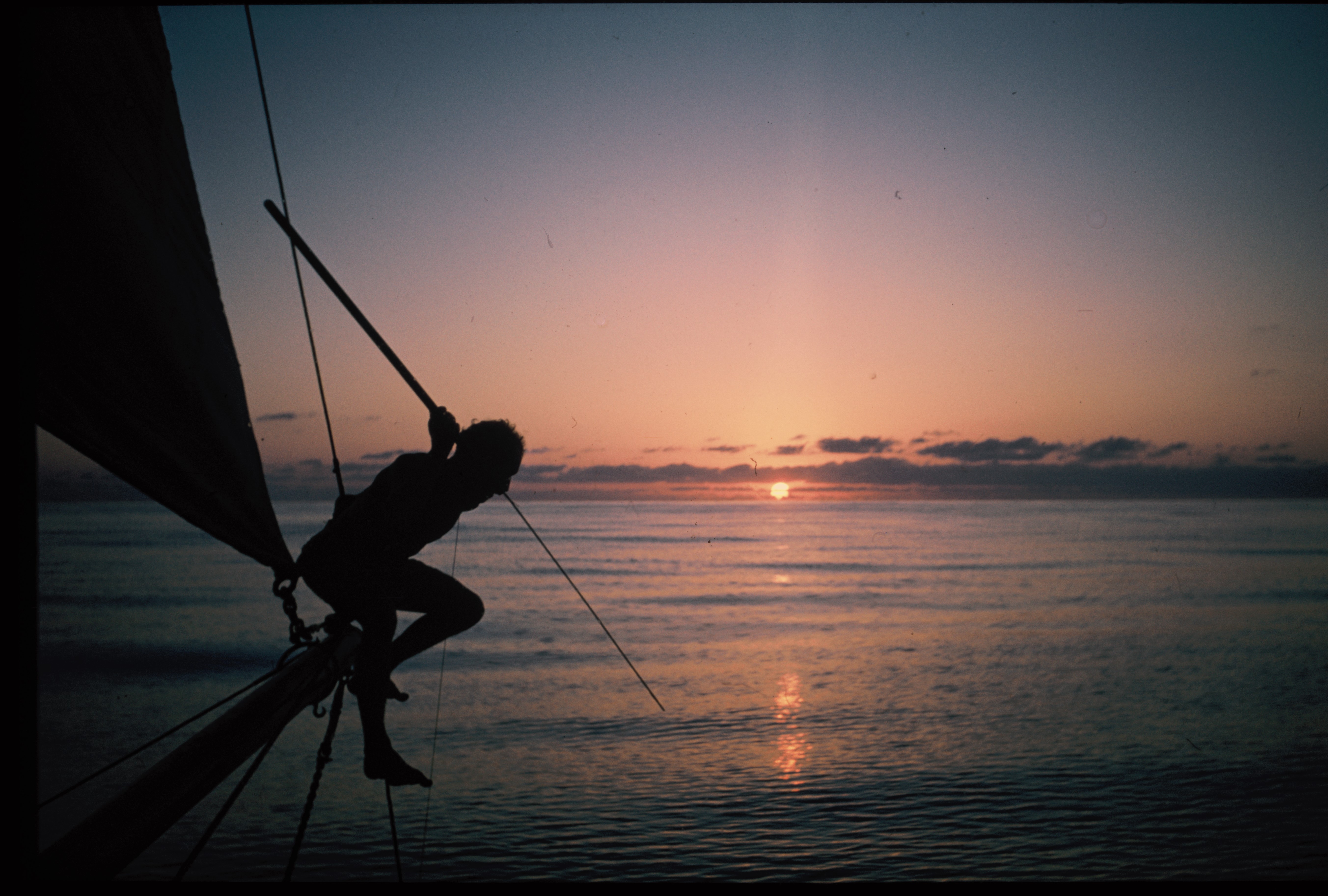En mann med fiskespyd sitter ytterst på en seilbåt i solnedgang