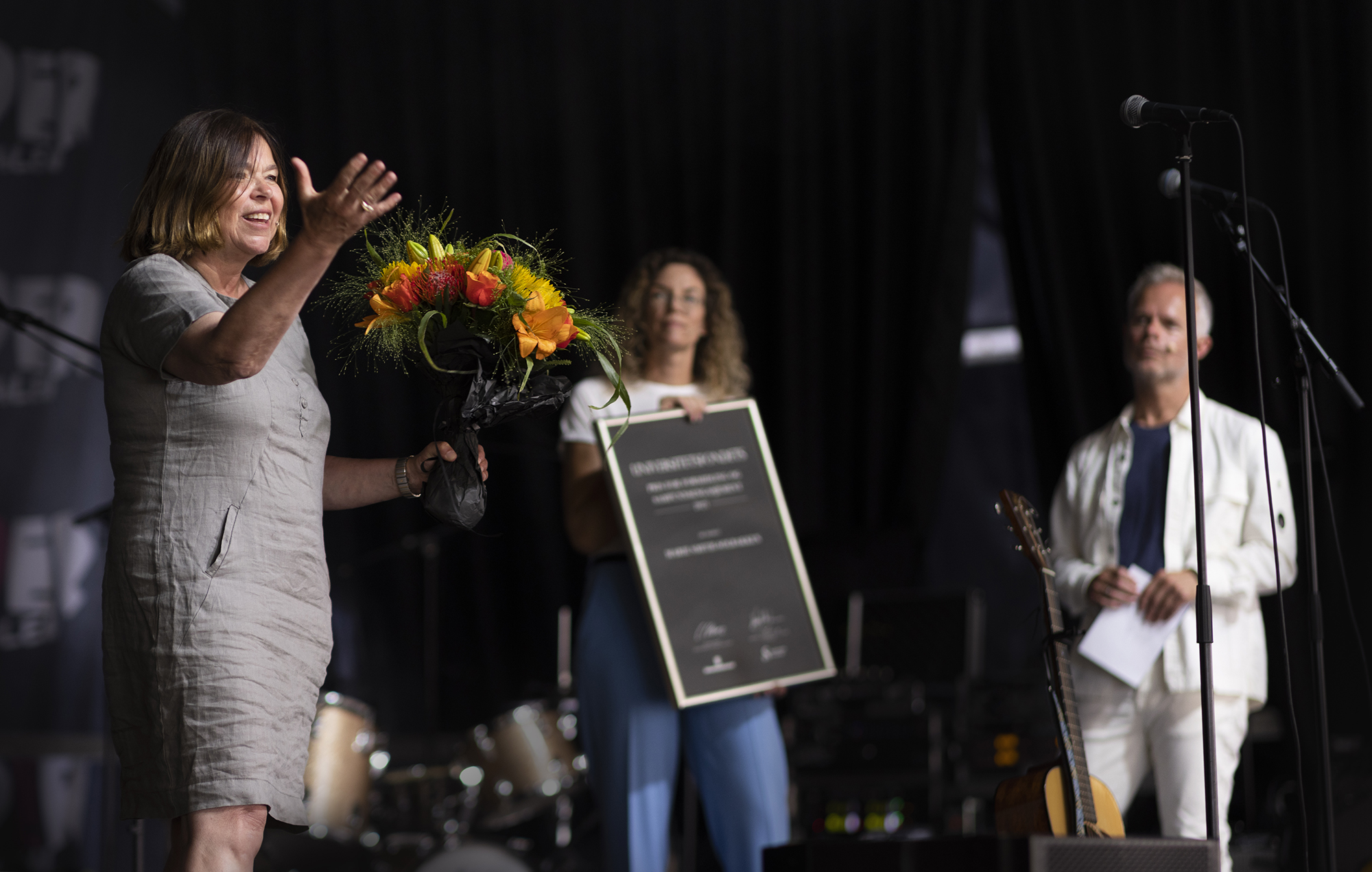 Prisvinner Marie Smith-Solbakken i grå kjole med blomster takker for pris. I bakgrunnen leder av Universitetsfondet, Cecilie Claviez som holder stort innrammet diplom, og lengst bak konferansier Tore Renberg.