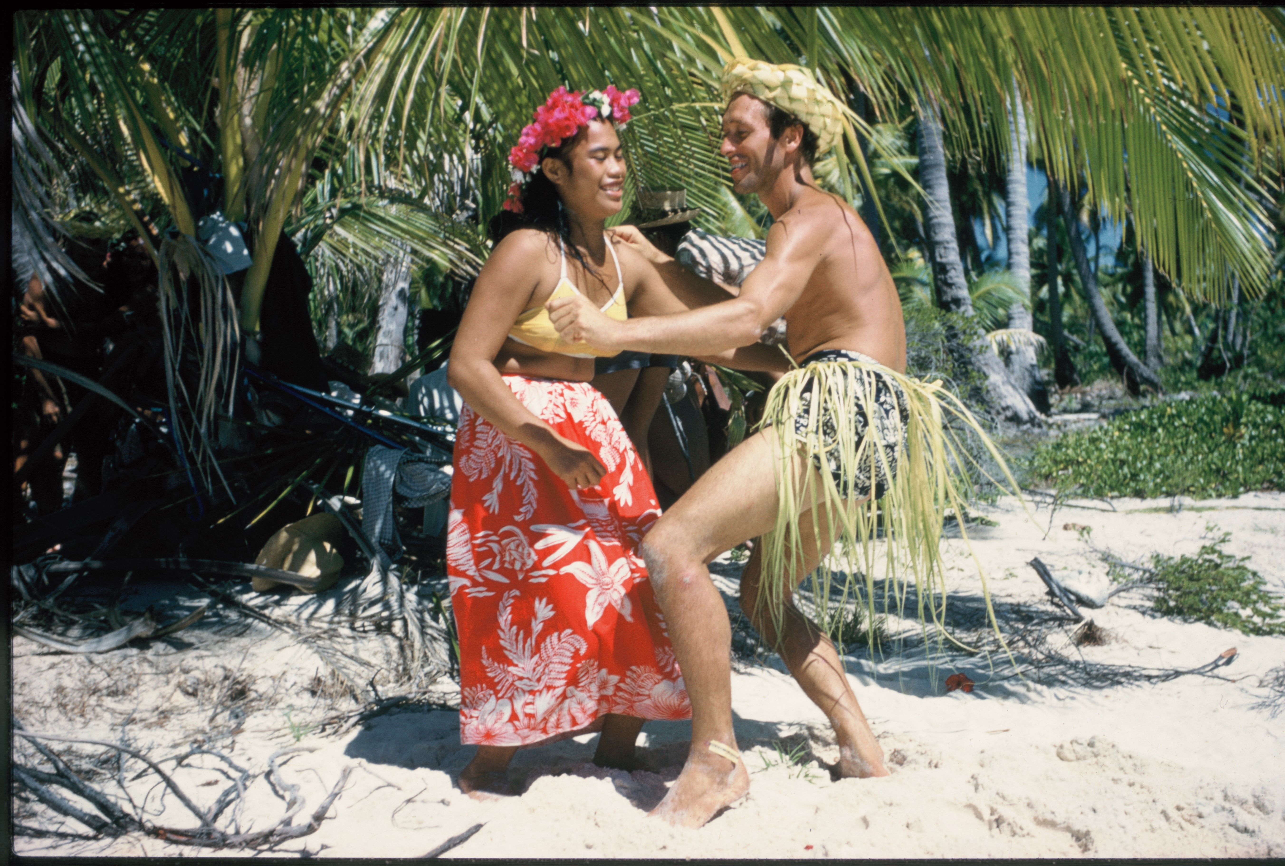 Et par urfolk med bastskjørt og blomsterkrans danser på en strand