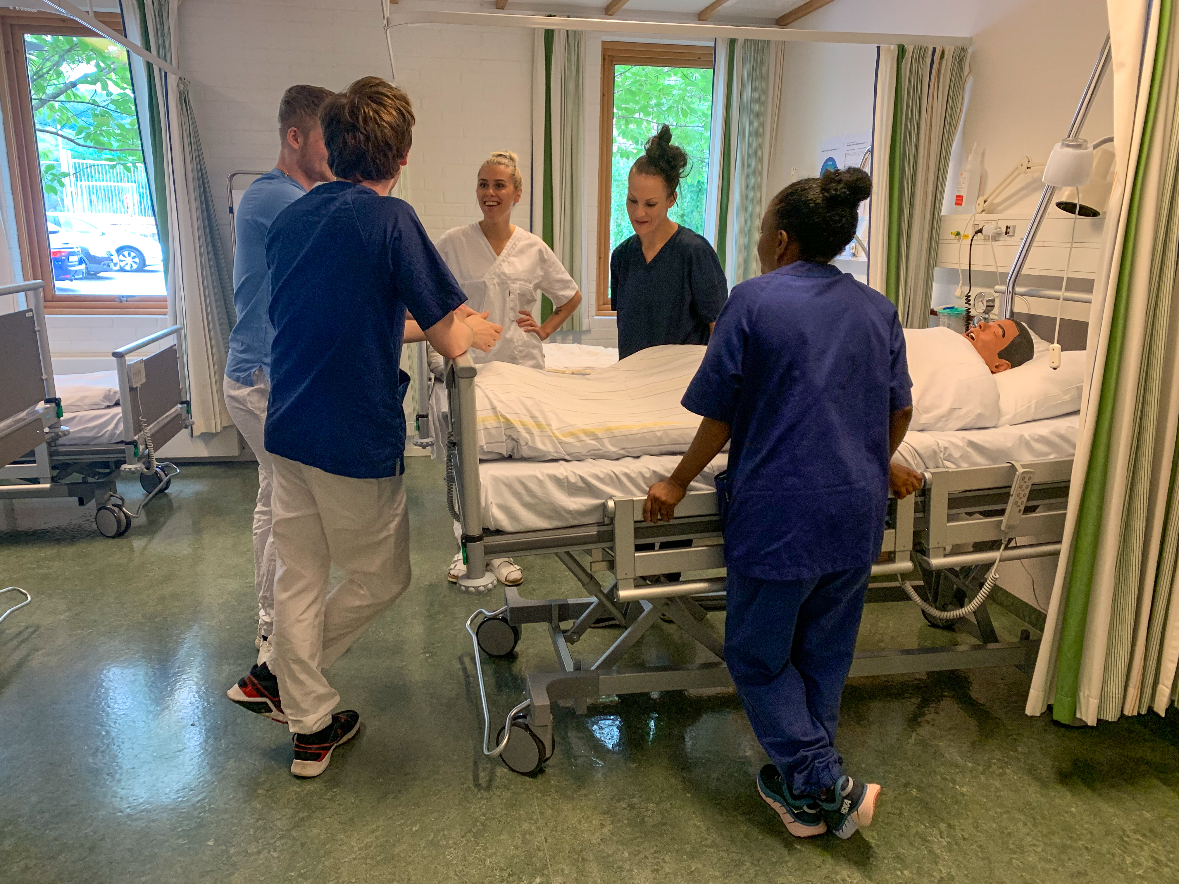 Fem sykepleierstudenter i uniform står rundt en sykehusseng under ferdighetstrening i lab