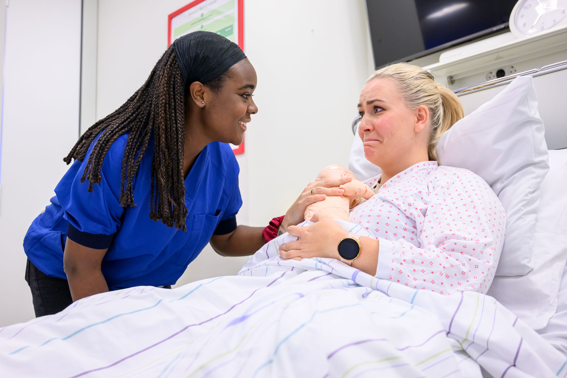 To jordmorstudenter under simulering. En student spiller jordmor og samtaler med student som spiller mor. Mor ligger i sykehusseng og holder en babydukke. 