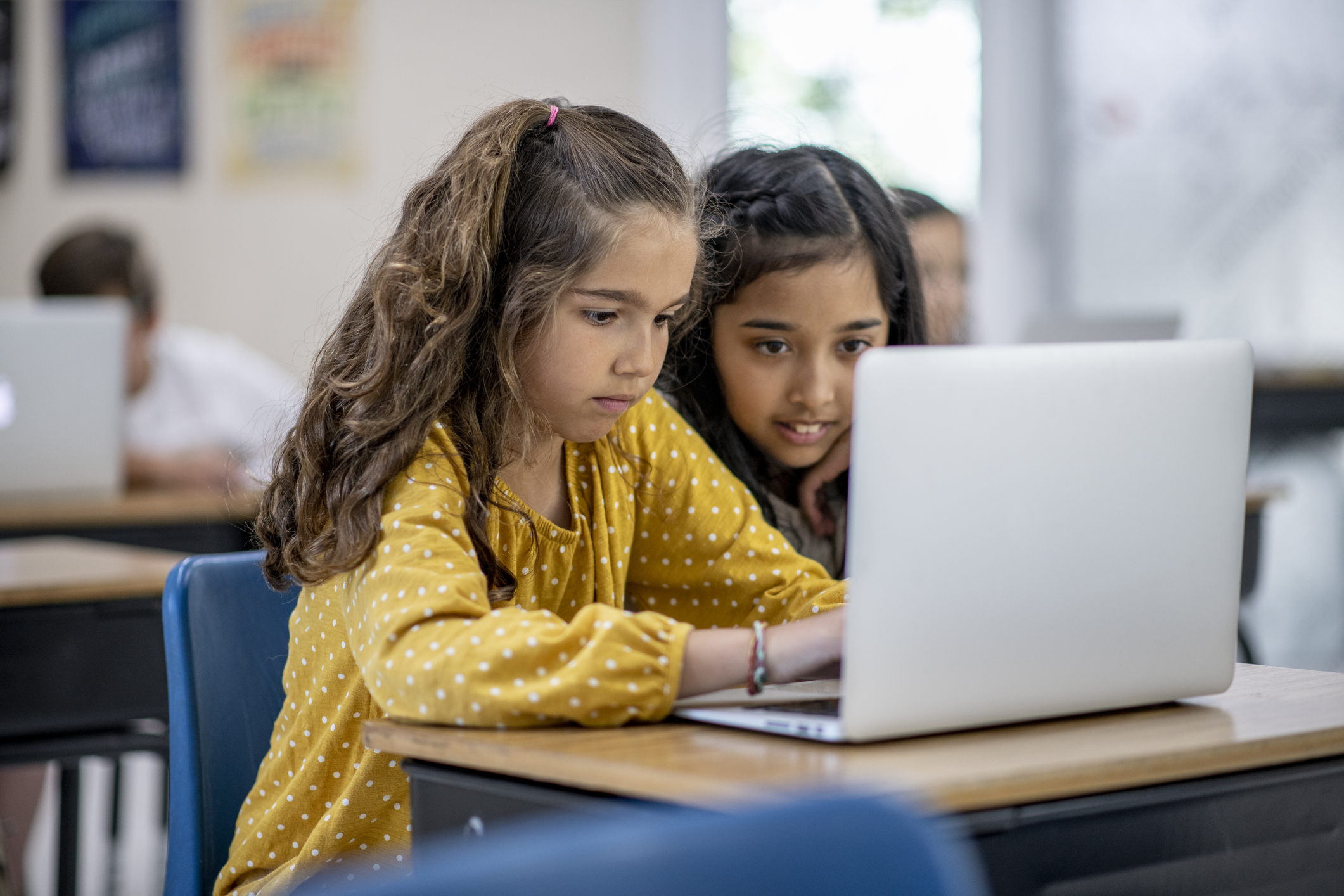 to jenter i barneskolealder sitter ved en pc. hun ene skriver, hun andre ser på og smiler til skjermen. 