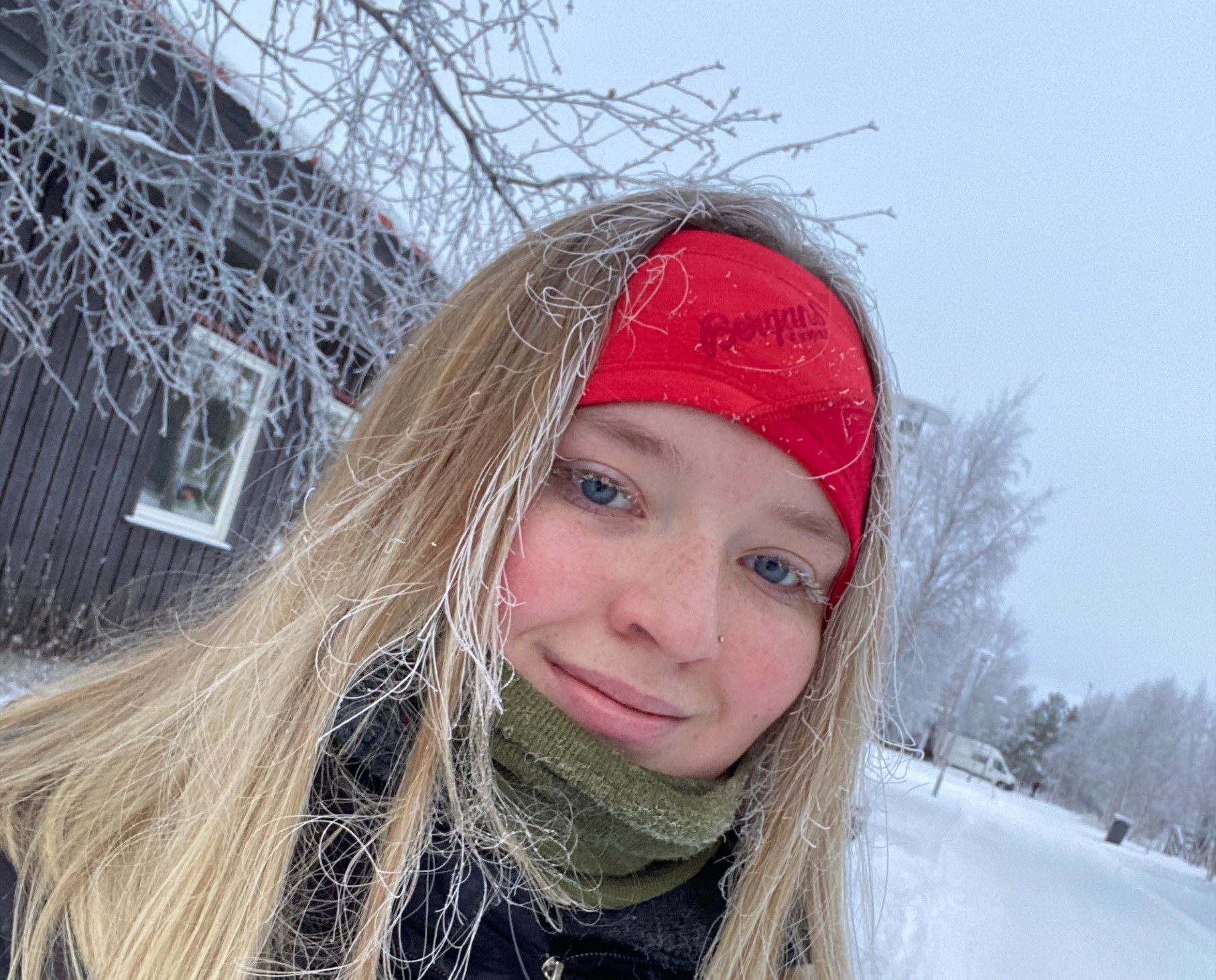 ung kvinne med langt, lyst hår og rødt pannebånd i vinterlig, hvitt landskap. Hun har snø i håret og frost på øyevippene.