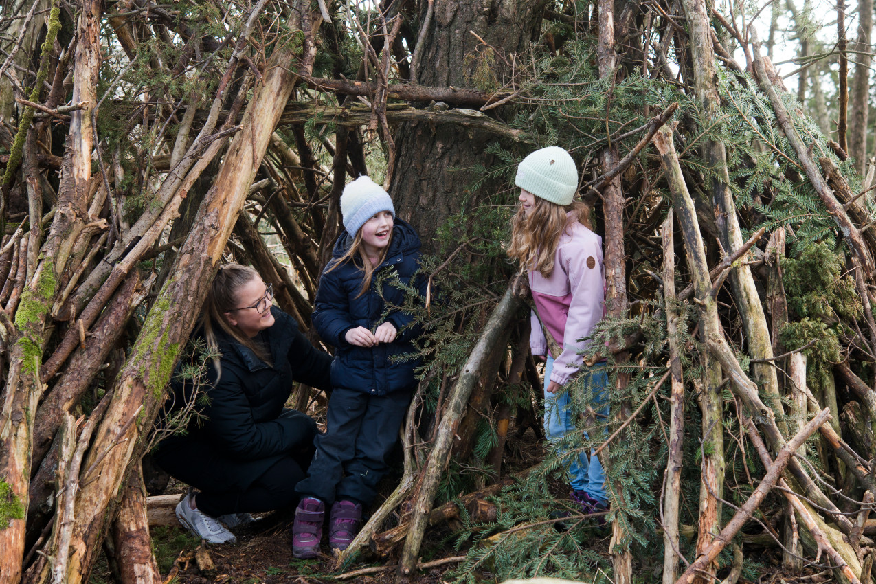 Lærer og to elever i åpningen av en lavvo laget av greiner og trær.