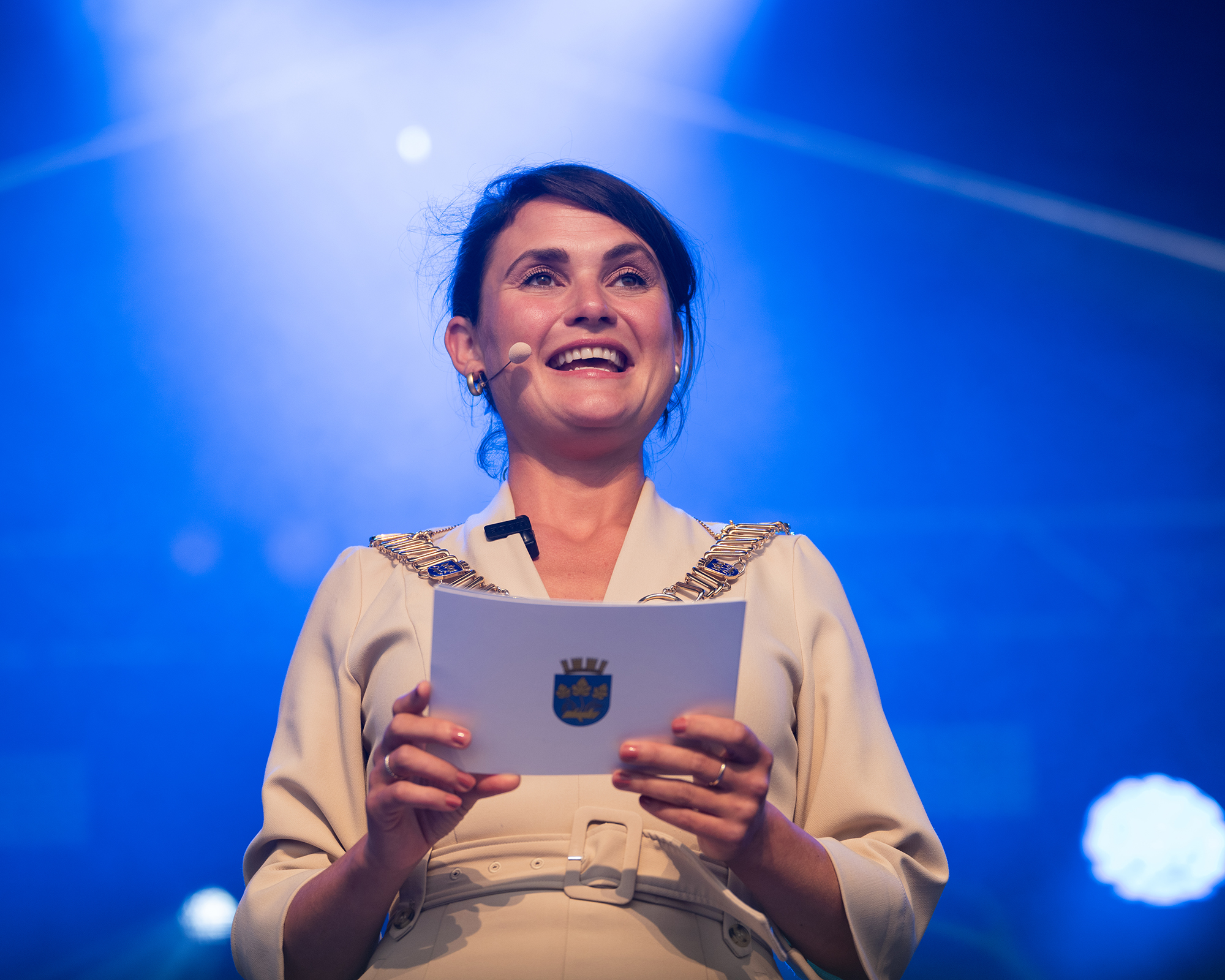 Kvinne med ordførerkjede og talekort med kommunevapen smiler fra scenen