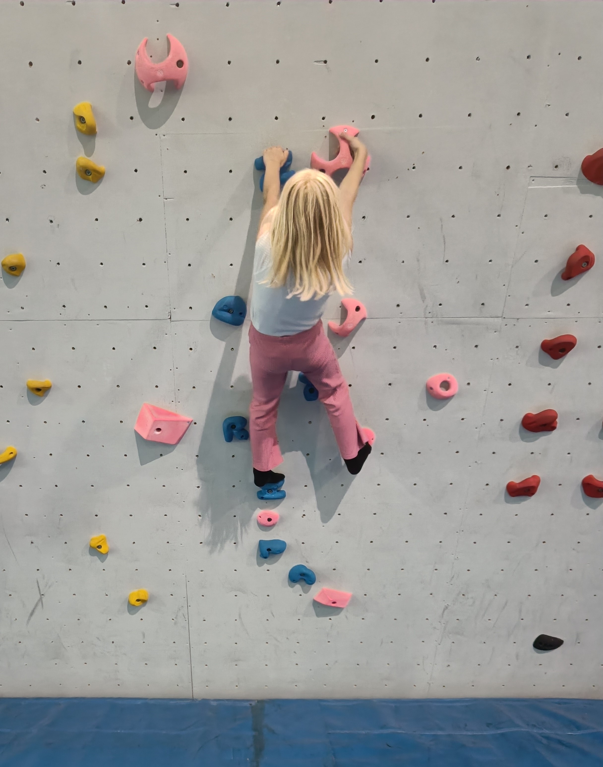 Et barn med langt, lyst hår, hvite og rosa klær har ryggen til kamera mens hun klatrer i en klatrevegg