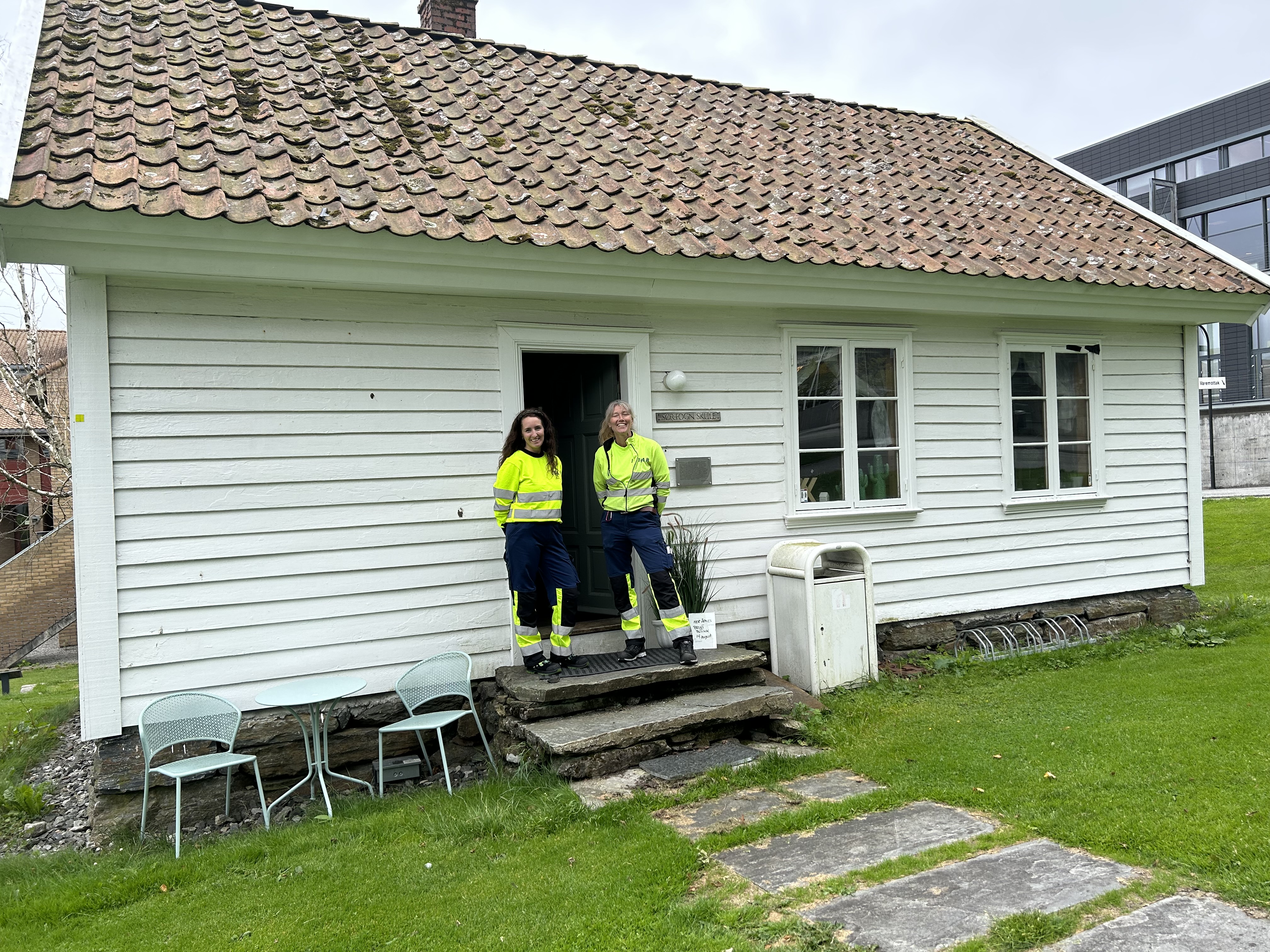 To kvinner i gul og blå dress står på trapp i inngangspartiet i lite trehus