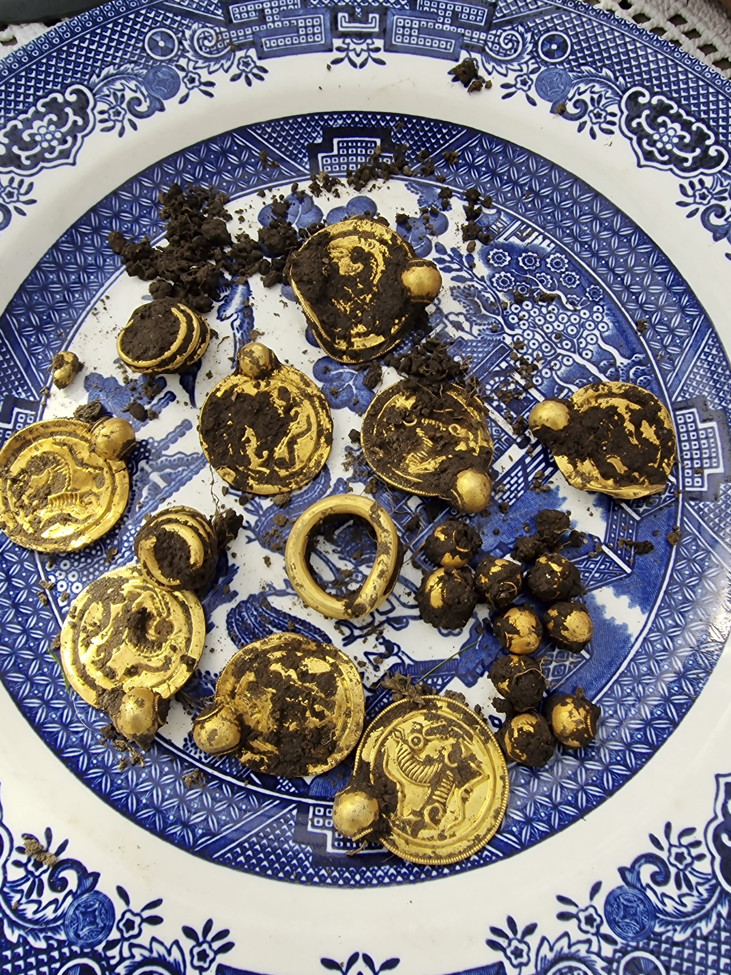 Gullskatt bestående av gullmedaljonger, gullperler og gullringer iblandet jord på en blåmønstret porselenstallerken.