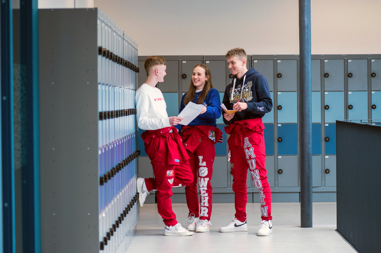 Tre elever, to gutter og en jente i midten, i russekklær prater smilende i en gang på skolen med skaper i bakgrunnen.