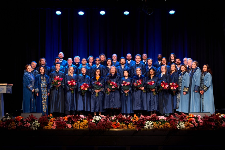 En stor gruppe mennesker oppstilt i blå frakker samlet på en scene og holder blomsterbuketter i hånden