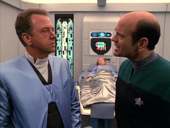 Skjermdump fra Star Trek Voyager episoden Critical Care. Viser to menn foran i bildet som diskuterer, med en pasient liggende i en seng i bakgrunnen.