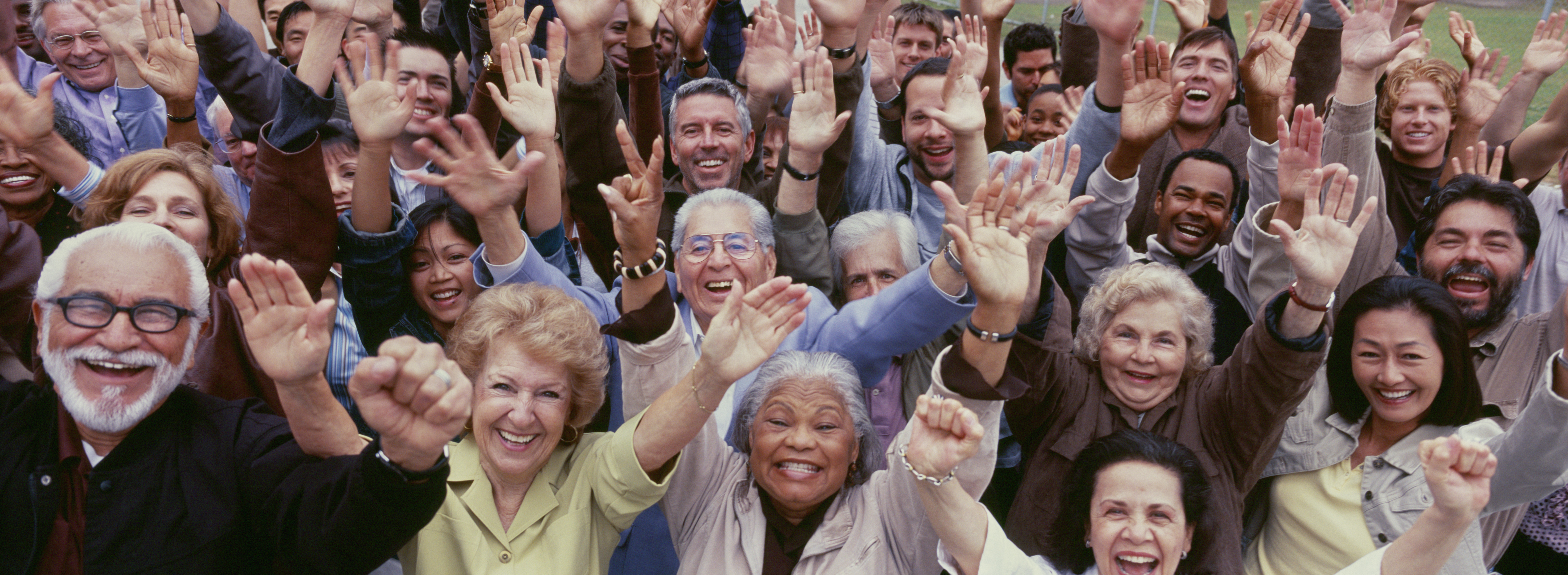 En stor gruppe blide voksne med forskjellig alder og etnisitet strekker hendene i været.