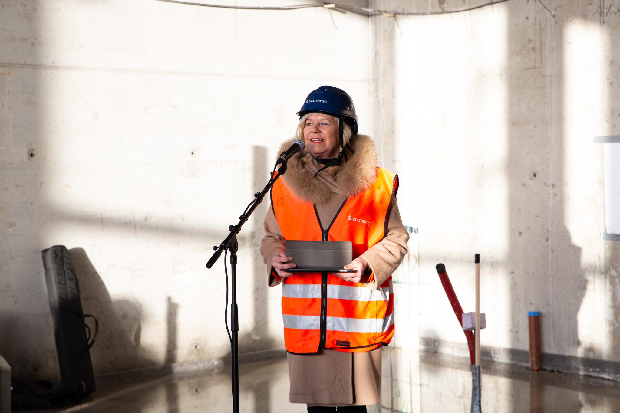 Kvinne med hjelm og refleksvest ved mikrofon i nybygg