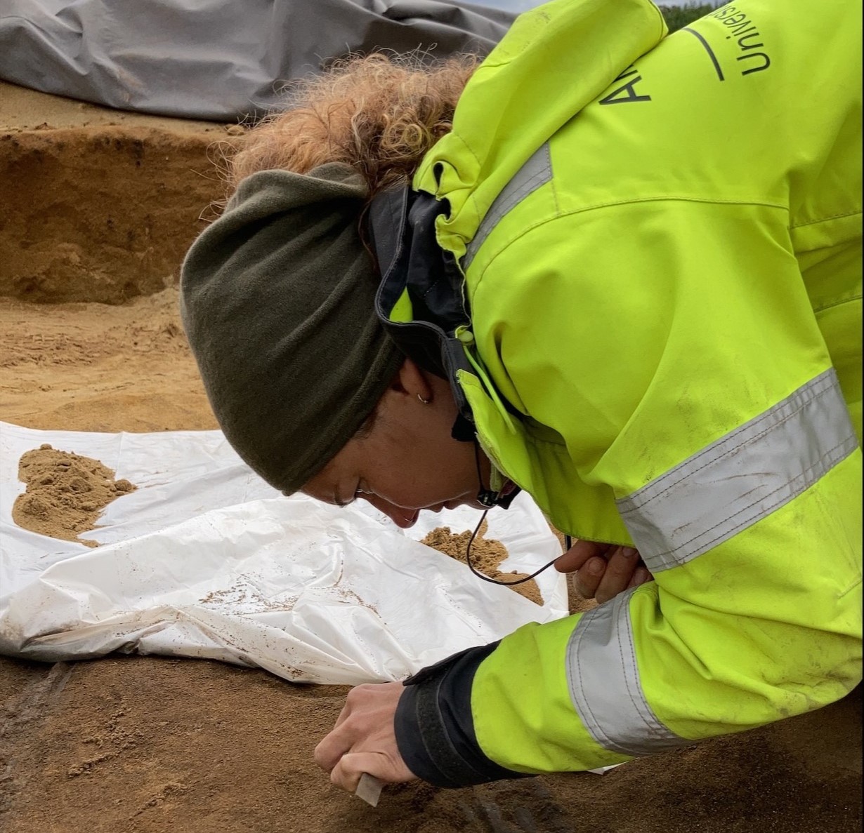 Arkeolog ligger og graver i jorda ved et gravfunn med et lite redskap. 