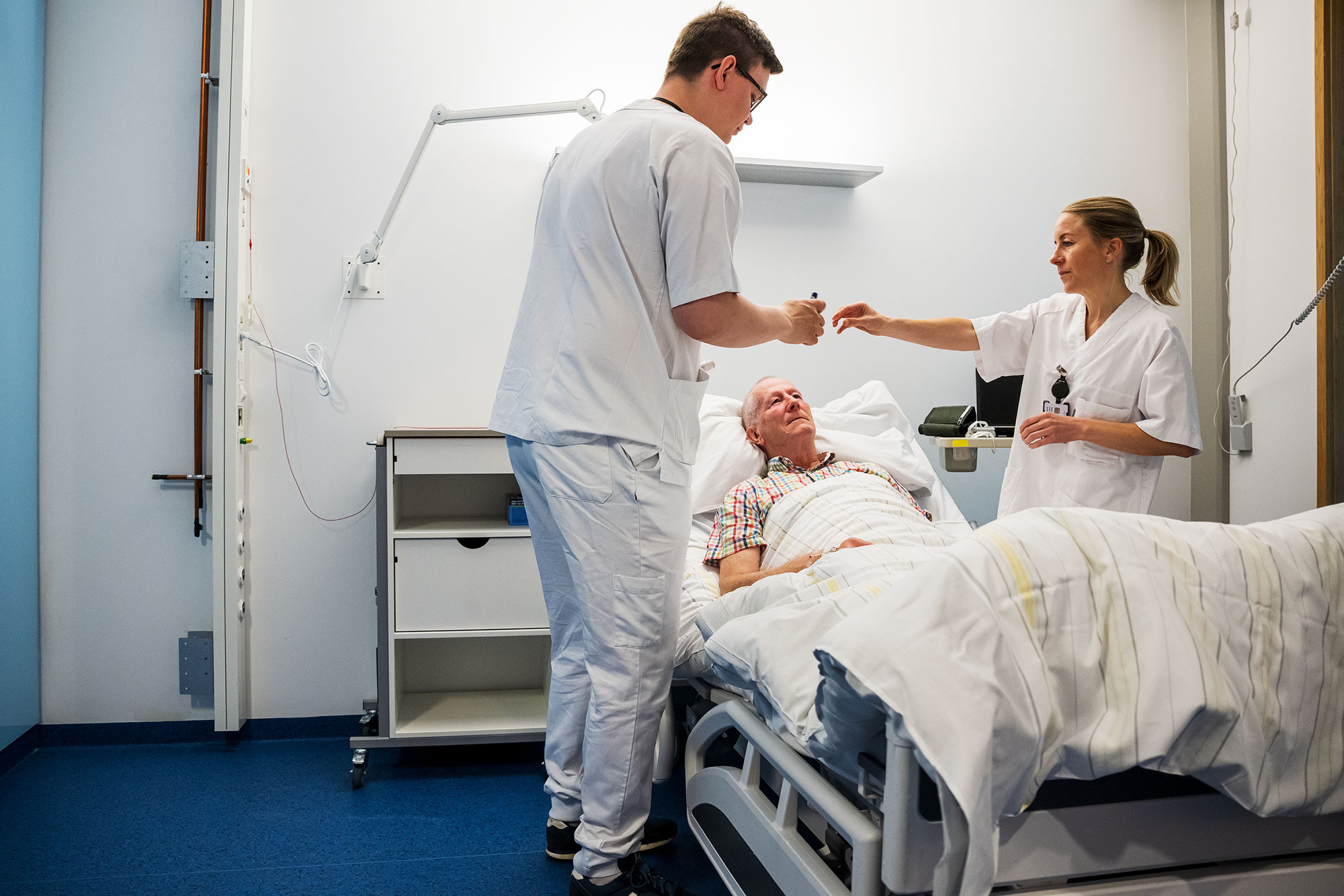 To sykepleiere står på hver side av en pasientseng på sykehus. Det ligger en eldre pasient i sengen.