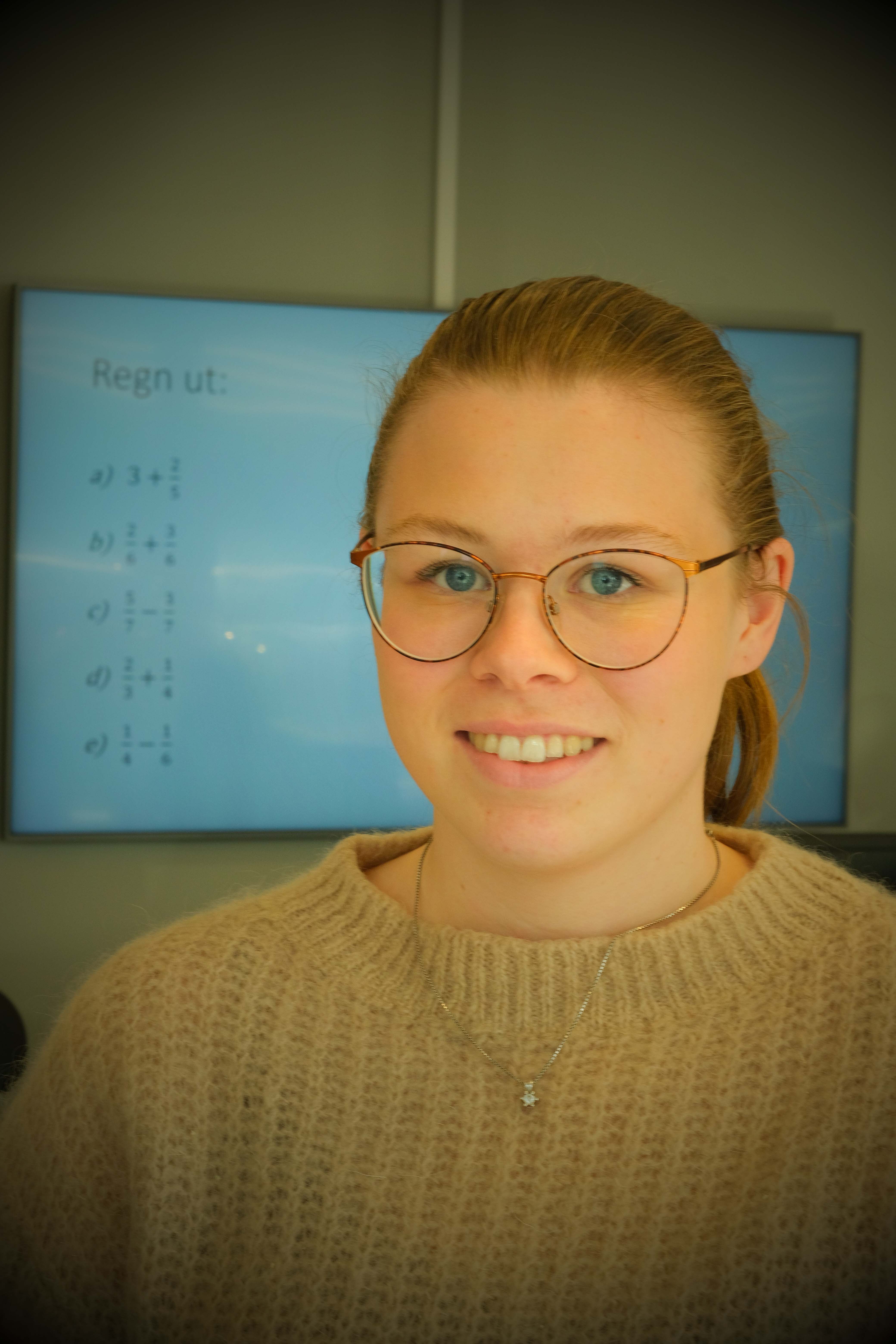 ung kvinne med håret i en hestehale smiler til kamera. Vi ser en tavle med matematikkstykker i bakgrunnen. 