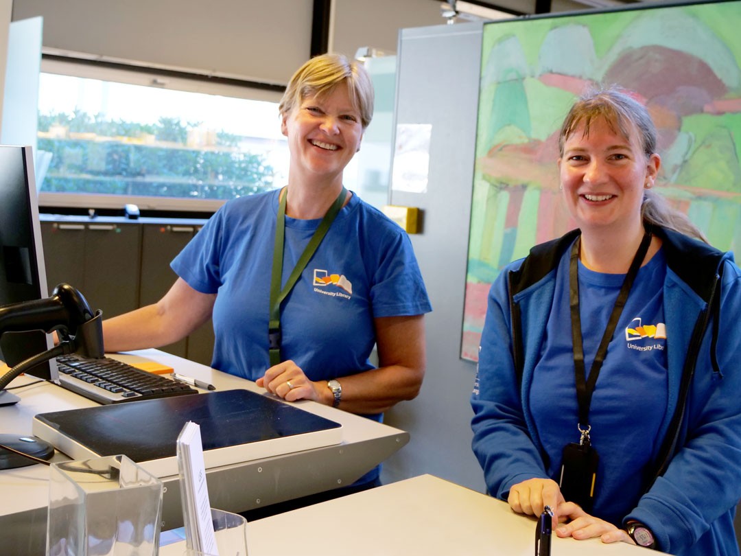 to smilende damer i blå trøyer står bak en serviceskranke 