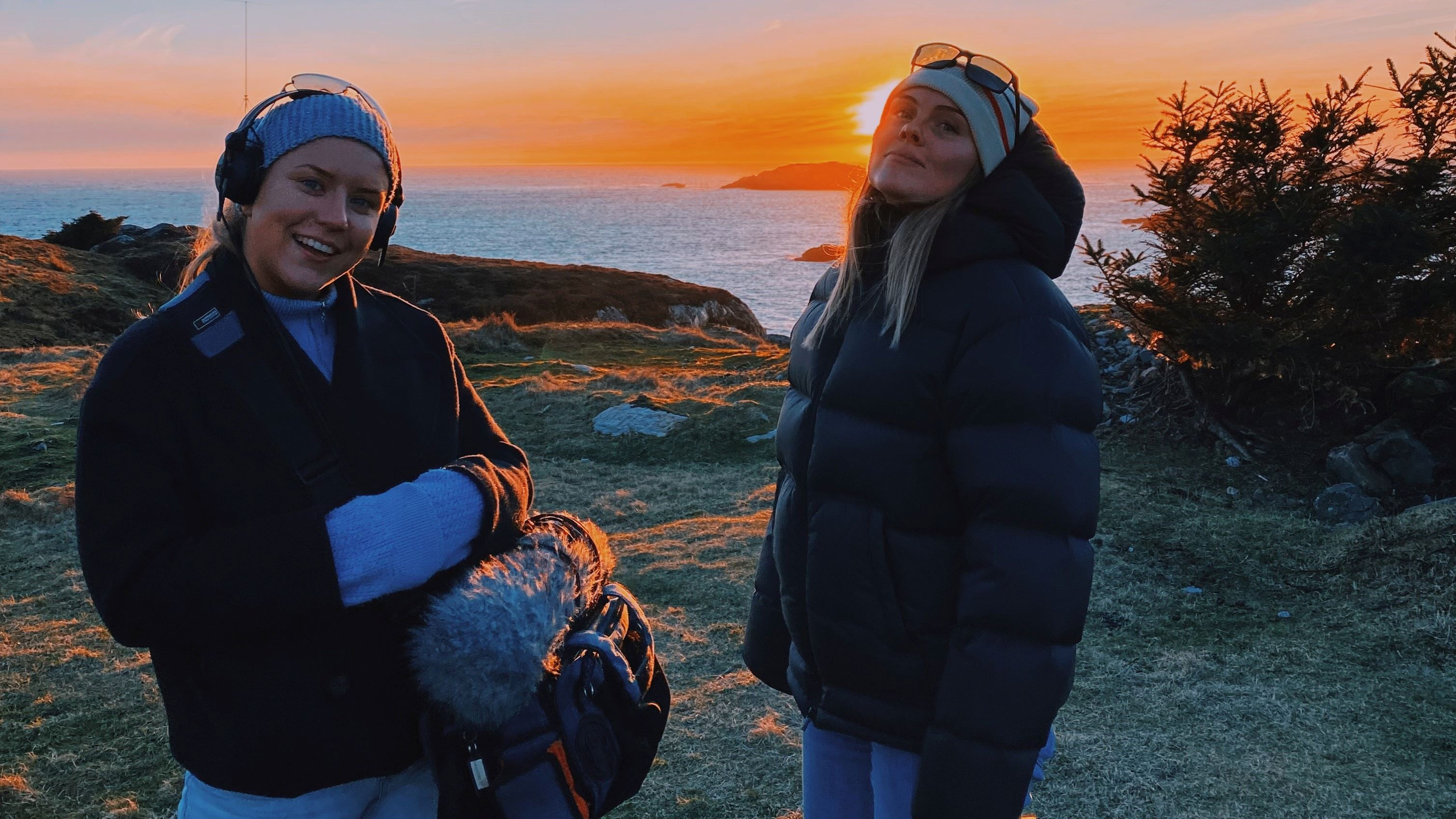 To unge kvinner iført dongeribukse og sort boblejakker står på en gresslette med havet og solnedgangen i bakgrunnen. Kvinnen til venstre holder et videokamera i hendene, og begge ser inn i kamera.