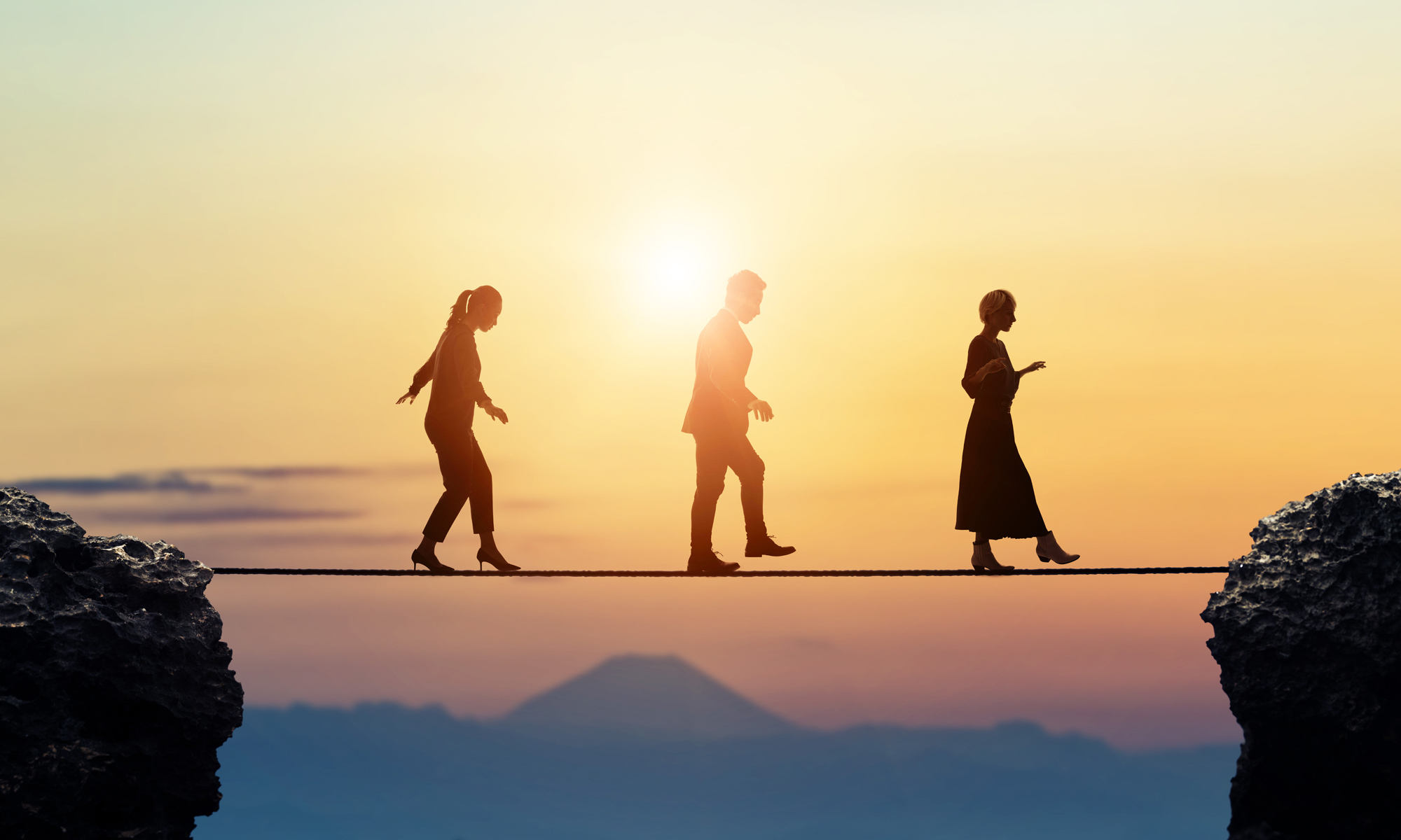 Business executives walking a tightrope over a ravine with mountains and sunset in the background.
