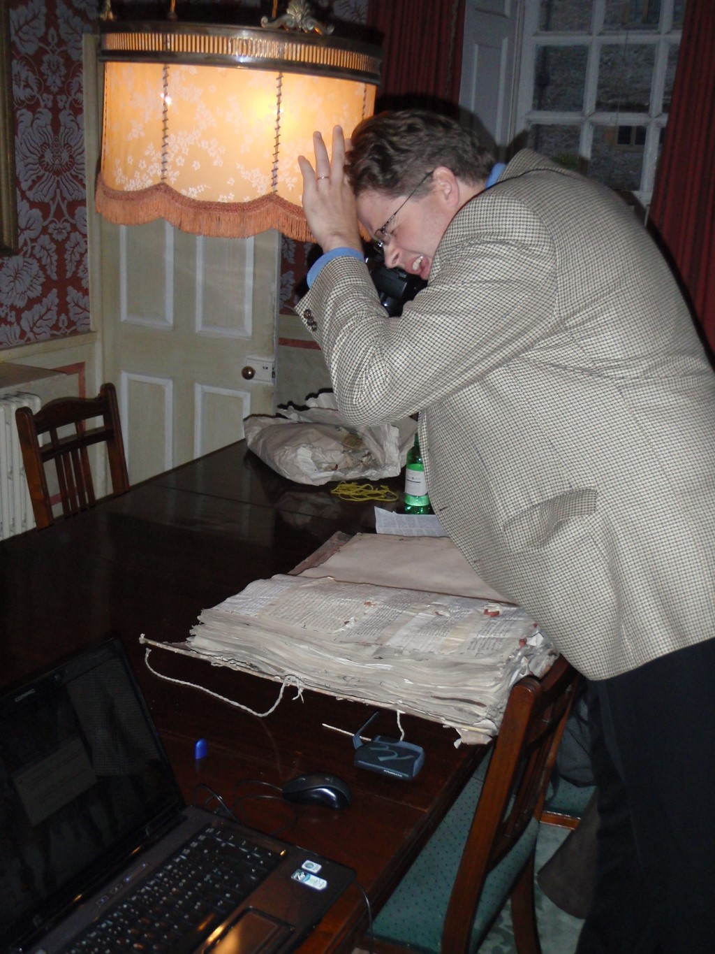 A man examines an ancient book in a study. The book is open on the table and the man is leaning over it and pushing the lampshade over the table away from him so that he can get a better look