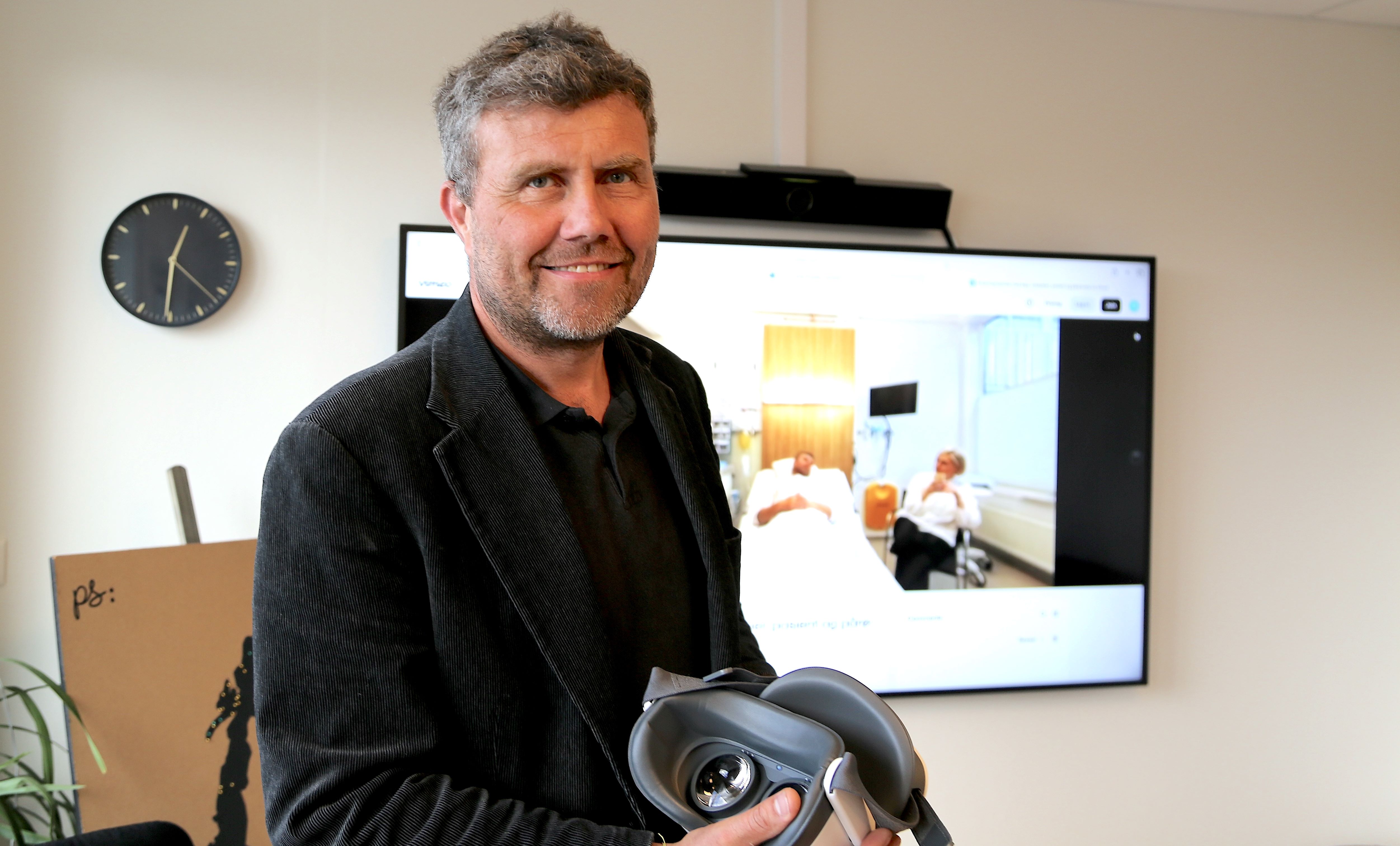 Mann med vr-briller i hendene og en skjerm bak seg