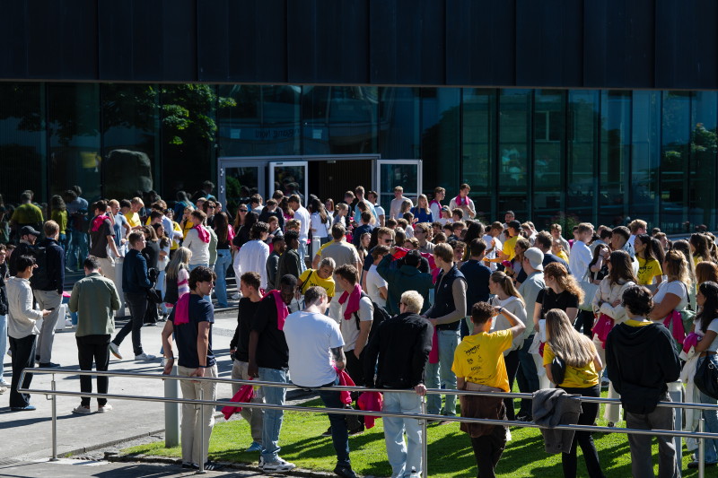 Mange studenter står tett i tett sammen i solen foran en mørk bygning.