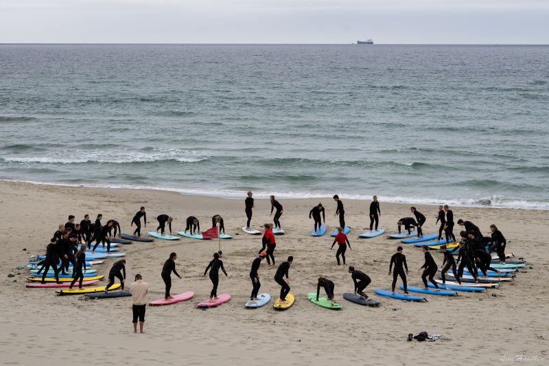 En gruppe ungdom i våtdrakt står på hvert sitt surfebrett i en ring på en sandstrand