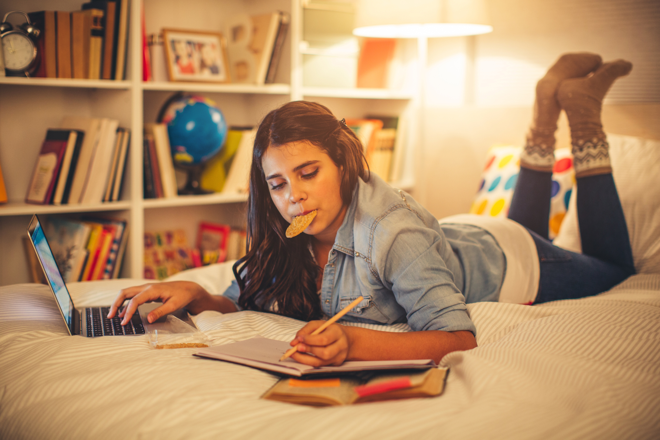 Jente som ligger på magen på senga med hånda på laptoppen mens hun skriver i en notatbok. Hun har en oppslått bok med postitlapper og en kjeks i munnen. 