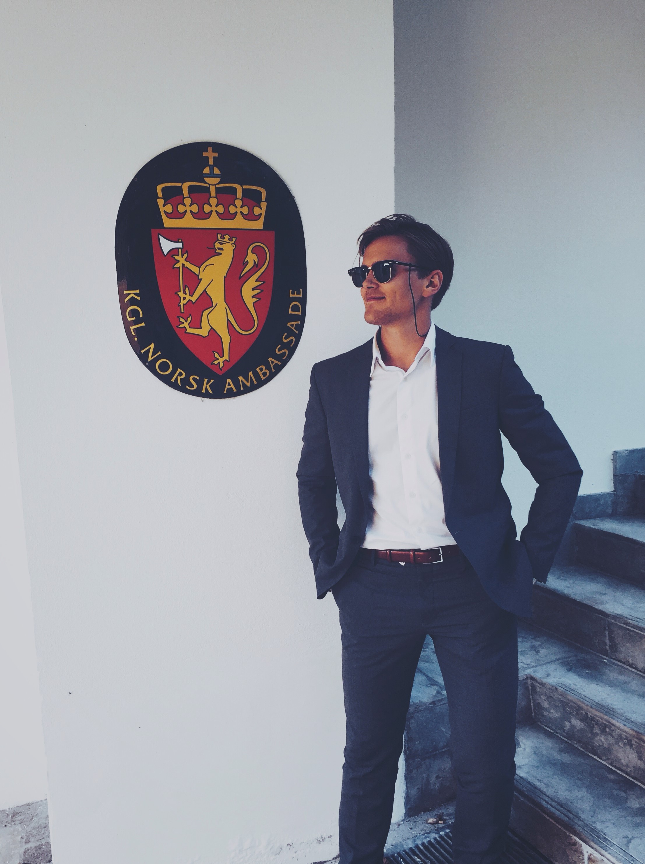 Med dress og solbriller inne i den norske ambassaden i København. Johannes står foran en vegg med bilde av det norske våpenskjoldet bak han: en sort oval med en rød trekant med krone oppå og en løve med en øks.