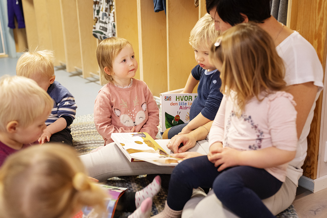 Barnehageansatt sitter i garderoben og leser bok for seks barn. Har god blikkontakt med et av barna. To andre barn sitter på fanget. De andre sitter rolig rundt henne i en sirkel.