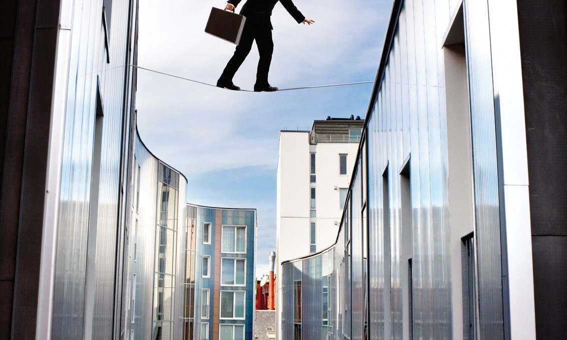 Mann i dress med stresskoffert går på stram line høyt mellom to bygninger