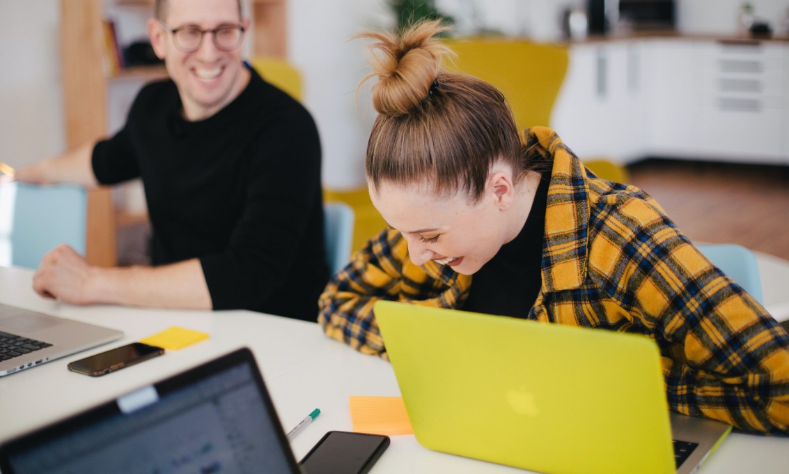 En kvinnelig og en mannlig student sitter med datamaskiner på et bord og ler sammen.