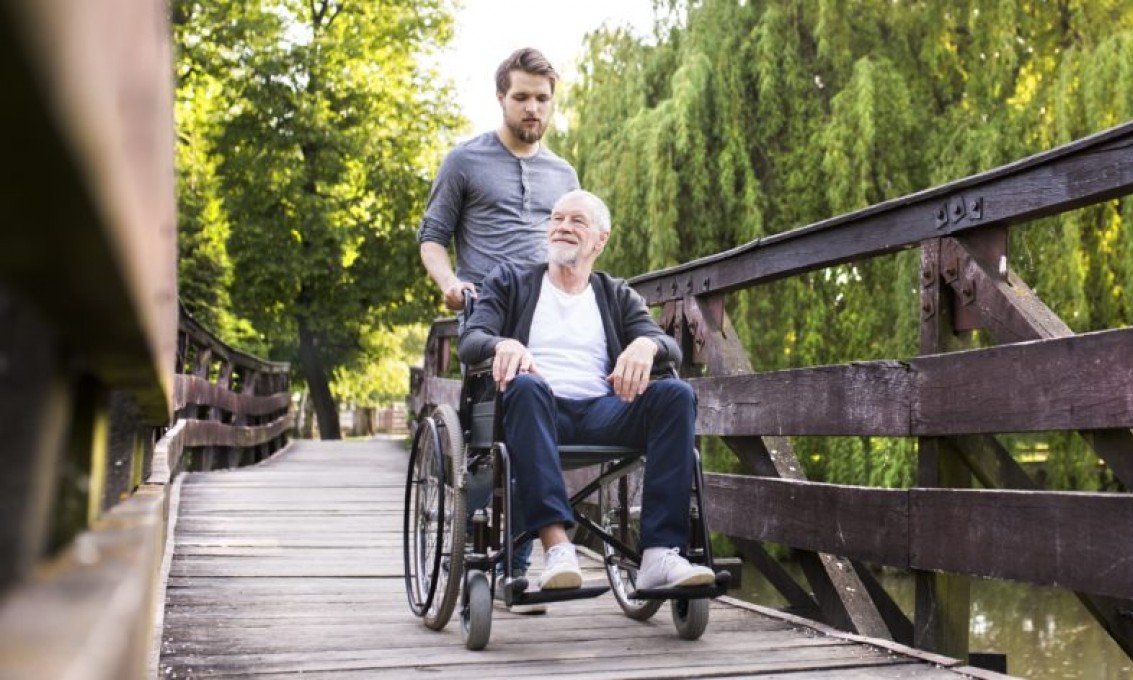 Sønn triller far i rullestol over en bro i en park