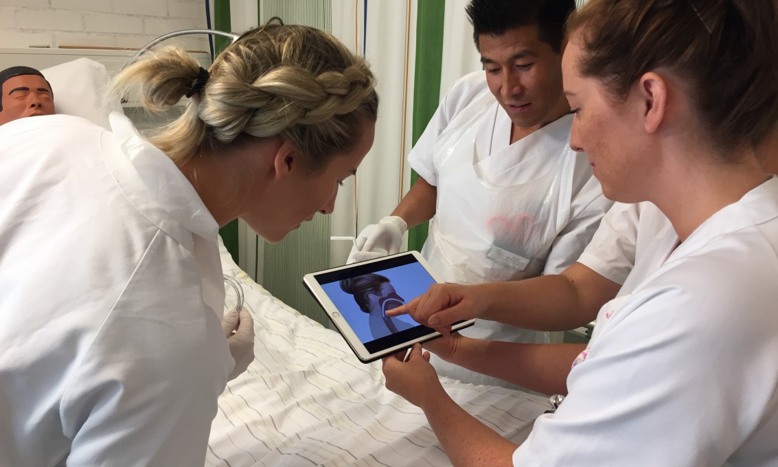 To kvinnelige og en mannlig sykepleierstudent i uniform ser en film på en ipad i sykepleielab