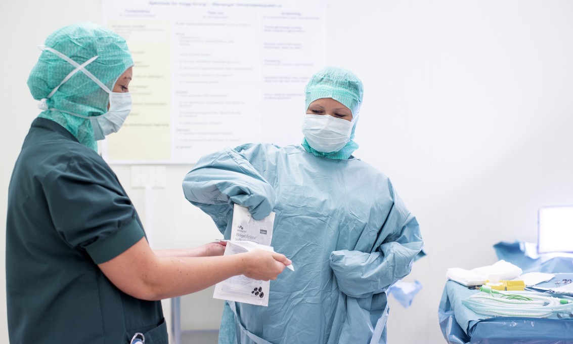 To kvinnelige operasjonssykepleiere i uniform på operasjonsstue. En av dem er sterilkledd og får assistanse til å ta på sterile hansker.