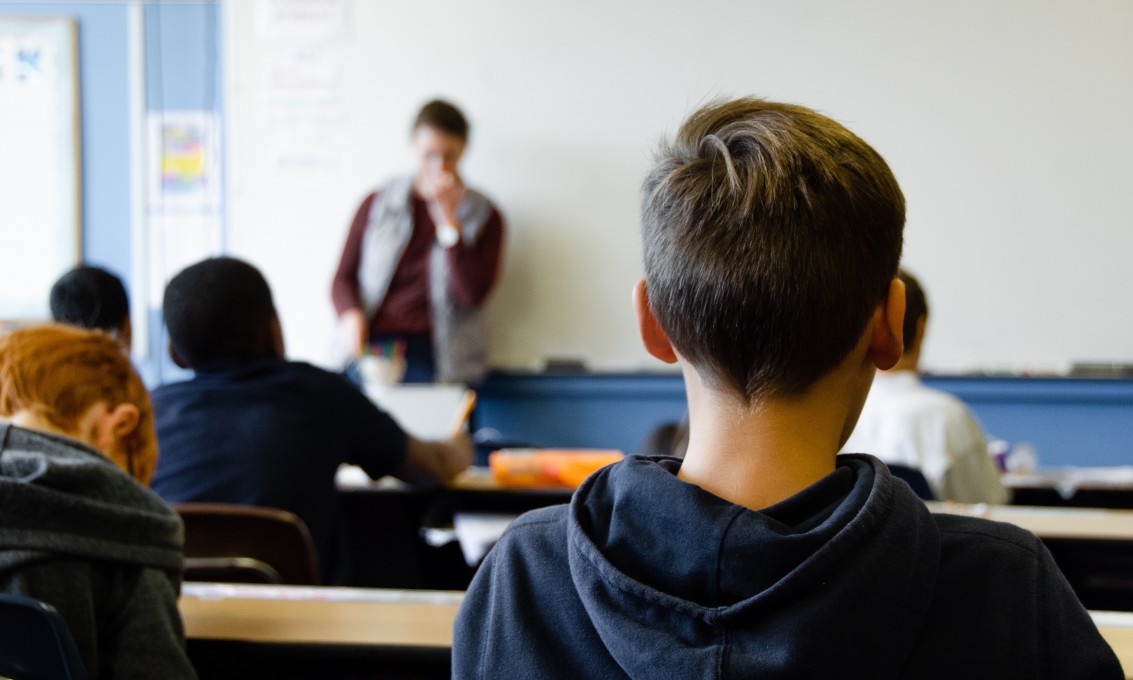 Bakhodet til en gutt. Lenger framme ser vi flere elever ved skolepulten og en lærer foran en hvit tavle. 