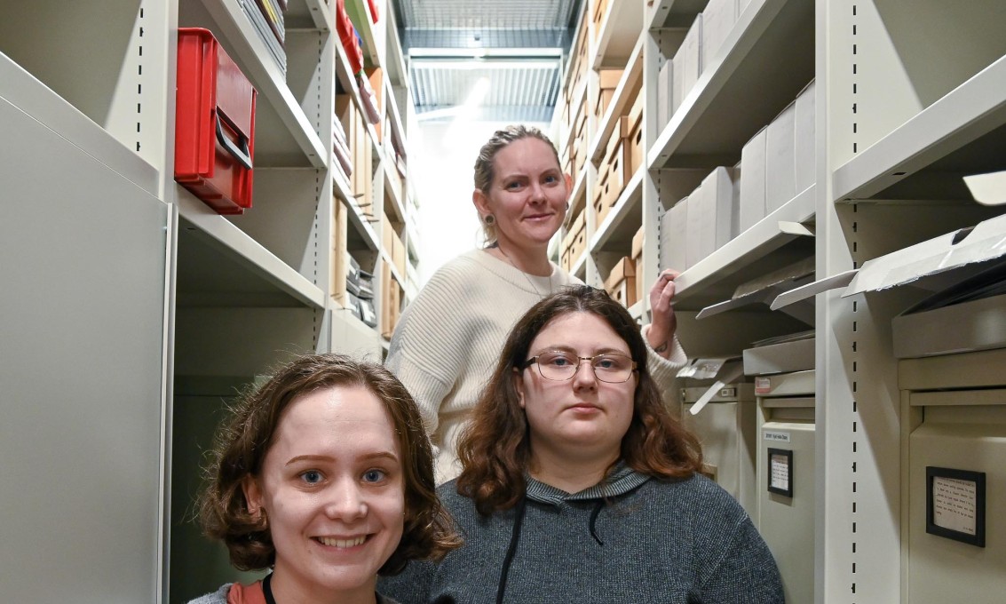 Tre unge kvinner står mellom to arkivhyller og ser rett i kamera