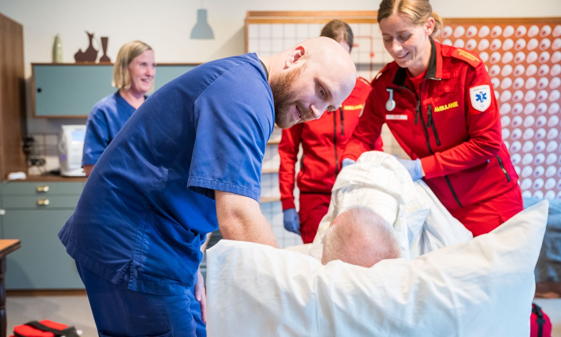 Mannlig sykepleier i blå uniform bøyer seg over pasient sett bakfra i en seng, mens kvinnelig paramedisiner i rød uniform hjelper til.