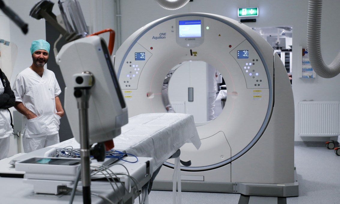 Hvit CT- og røntgenmaskin. En mann står på venstre side av maskinen. Foto. 