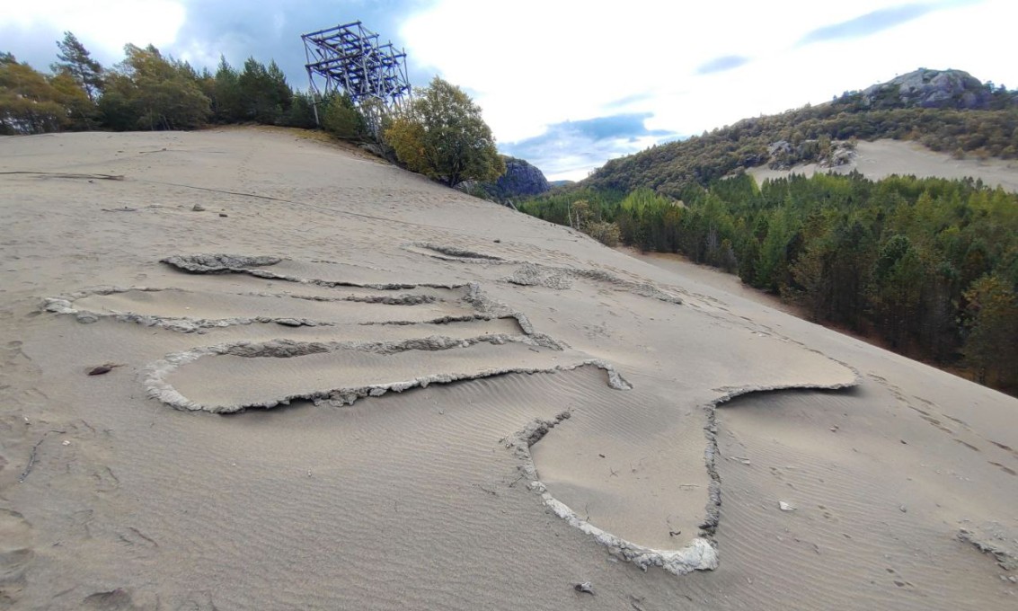 Bilde av et mønster i en sandbak. Et stativ, skog og himmel i bakgrunnen. Foto.