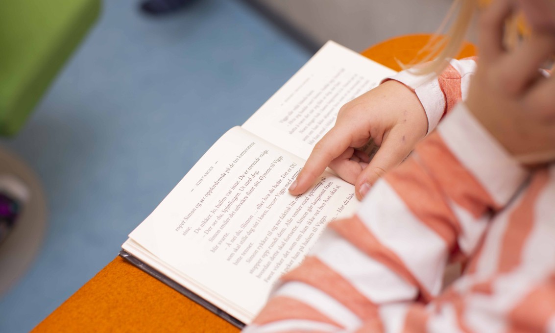 Bilde av en bok og utsnitt av et barn som leser. Ser bare hånden og genseren til barnet.