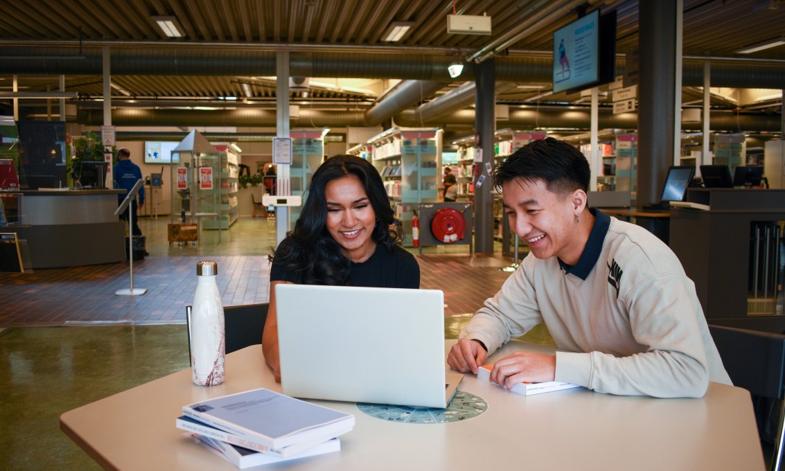 To studenter sitter sammen foran en laptop ved et skrivebord. Bibliotek i bakgrunnen.