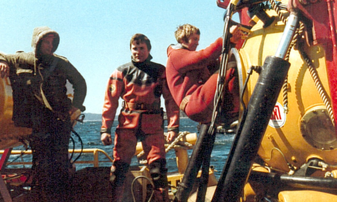 tre menn i dykkerdrakt på en oljeinstallasjon. En av dem er på vei inn i en dykkerklokke.