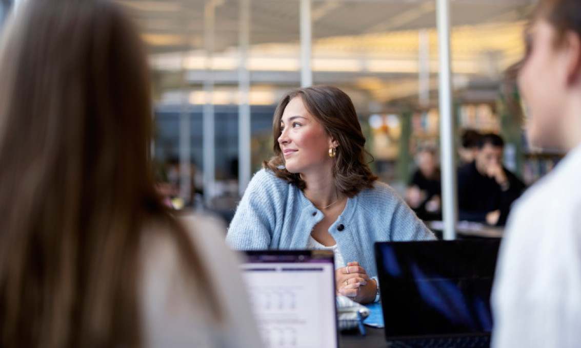Ung kvinne med skulderlangt brunt hår og lyseblå ullcardigan i arbeids- eller studiesituasjon er i fokus i midten av fotoet. Hun sitter vendt mot to andre som har ryggen mot kameraet.